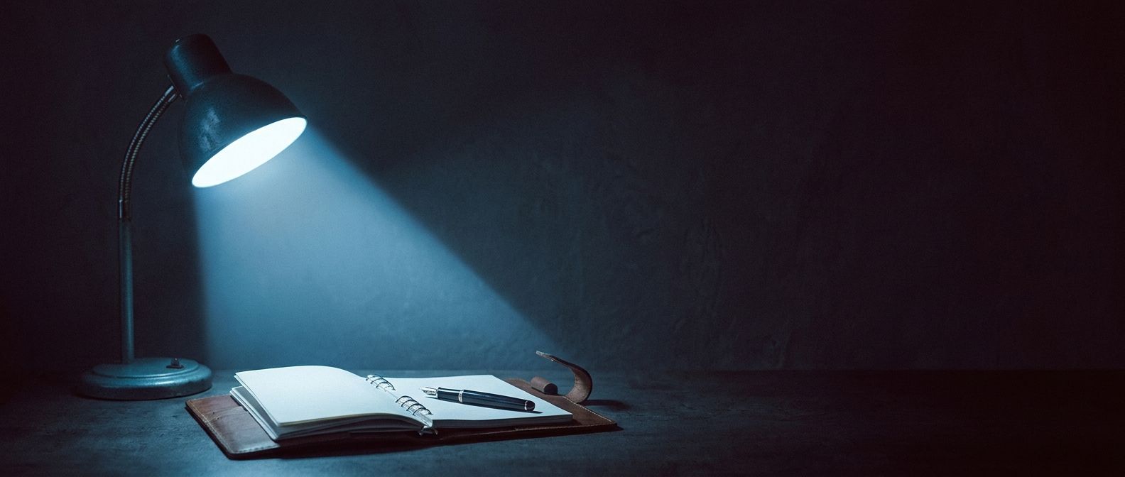 A desk lamp casting a single cone of light onto an open notebook in a dark room